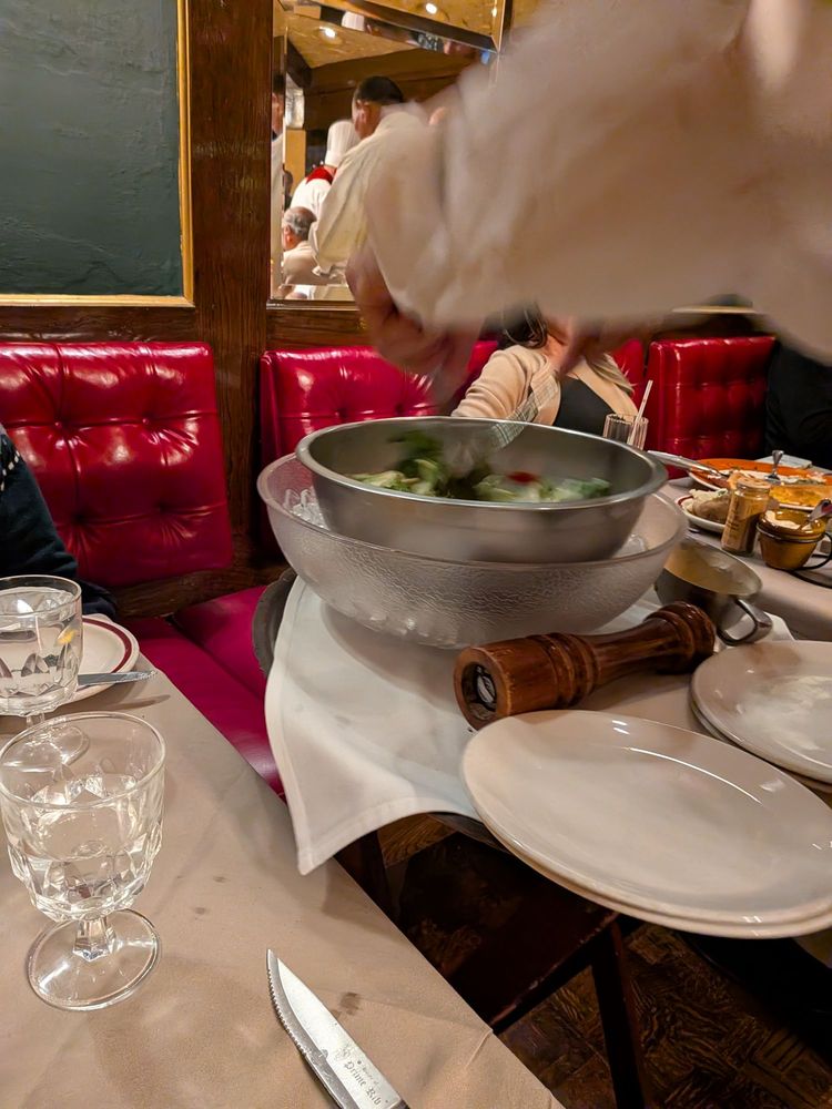 A server tosses salad tableside in a spinning bowl beside red tufted booths